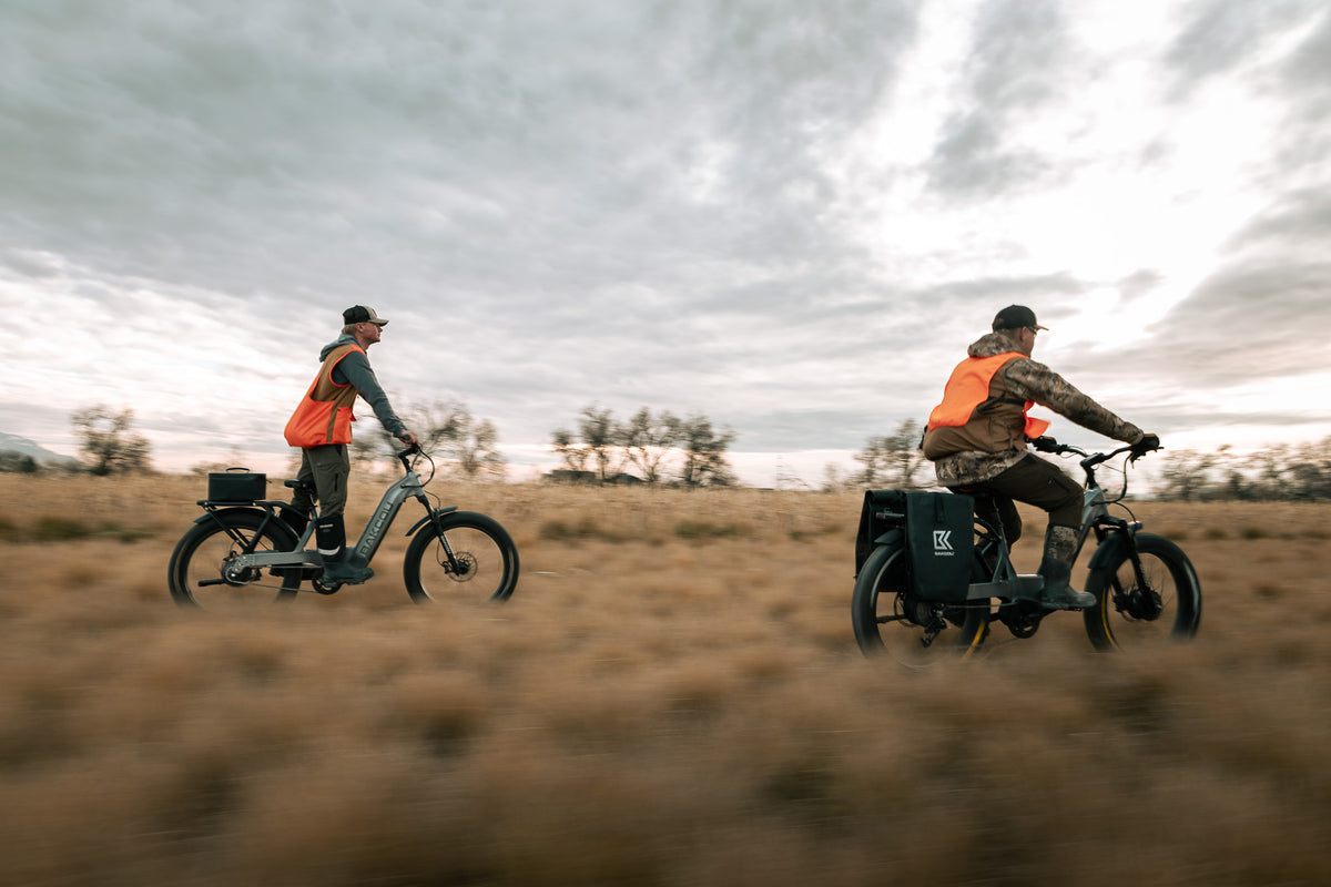 Two people riding electric bikes through a field wearing camouflage and hunter orange.