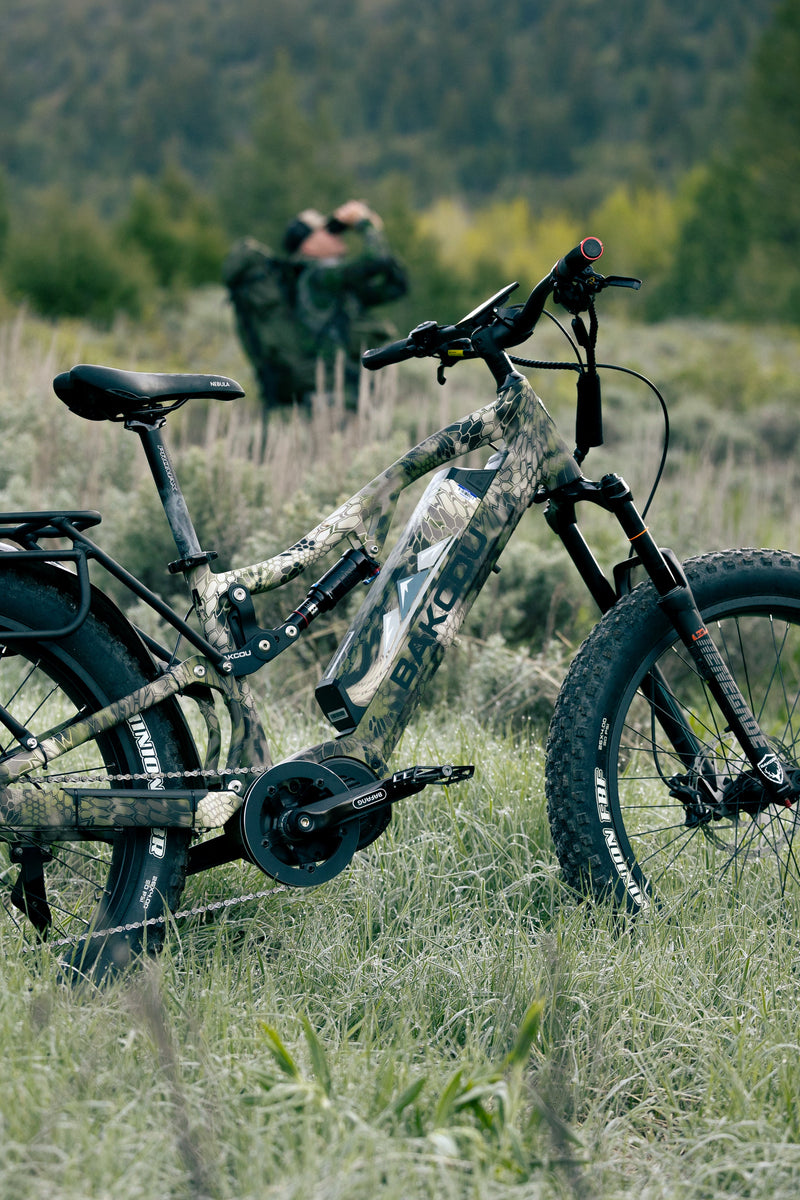 A fat-tire electric mountain bike with a camouflage pattern frame and rear rack, parked in a grassy field with trees in the background.