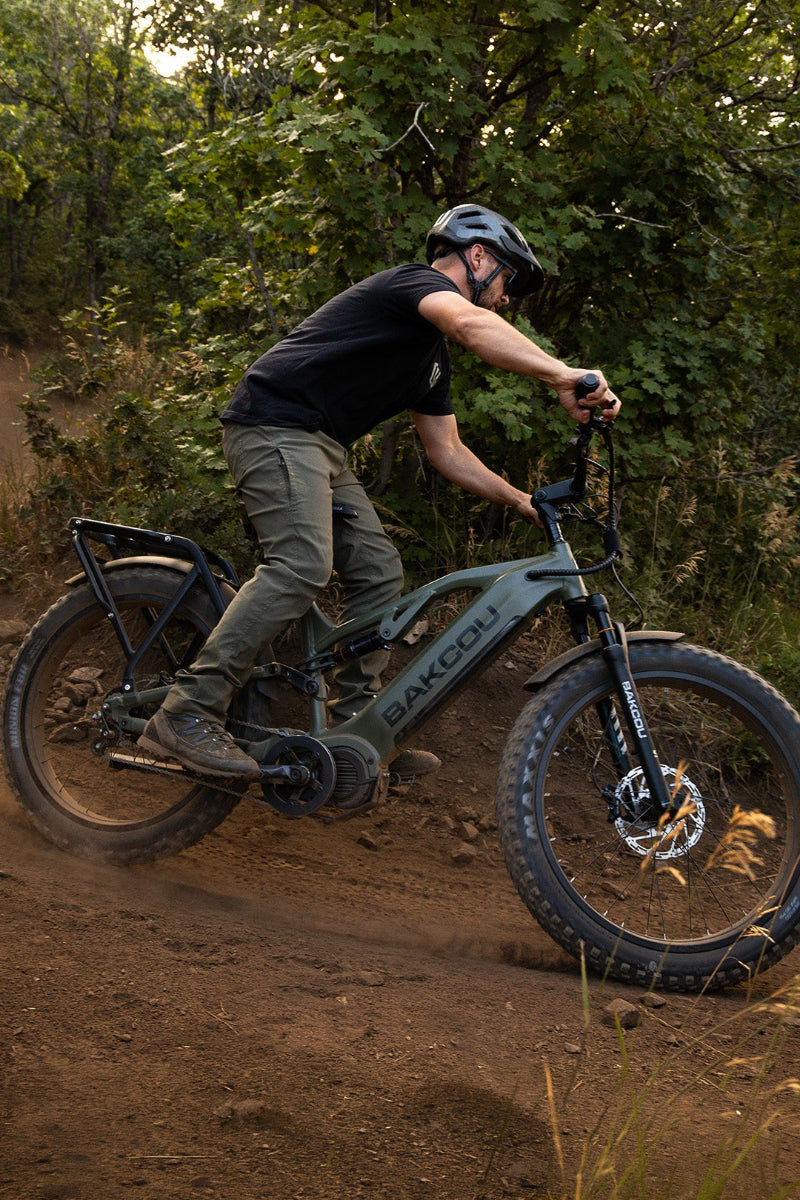 A man riding an electric fat-tire bicycle on a dirt trail through a wooded area. The bike frame is dark gray with the brand name 'BAKCOU' visible on the downtube.