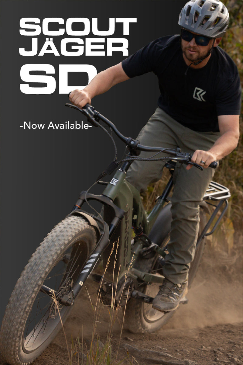 A man riding an electric mountain bike outdoors on a dirt trail, wearing a black Scout Jäger SD shirt, gray pants, and a silver helmet. The image features a dark gray graphic overlay on the left with the text 'SCOUT JÄGER SD -Now Available-'.